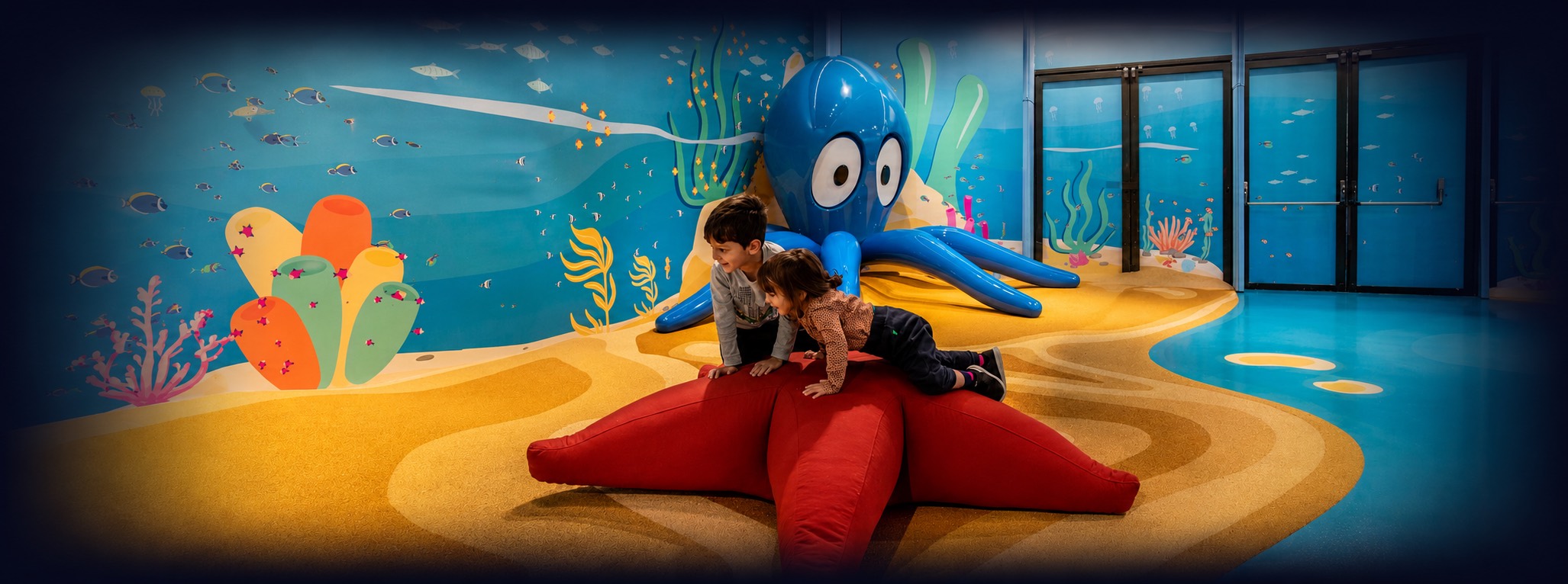Bambini giocano alla Città dei bambini e dei ragazzi Due bambini giocano su una grande stella marina rossa nell’area gioco a tema marino della Città dei Bambini e dei Ragazzi di Genova, con un grande polpo blu sullo sfondo.